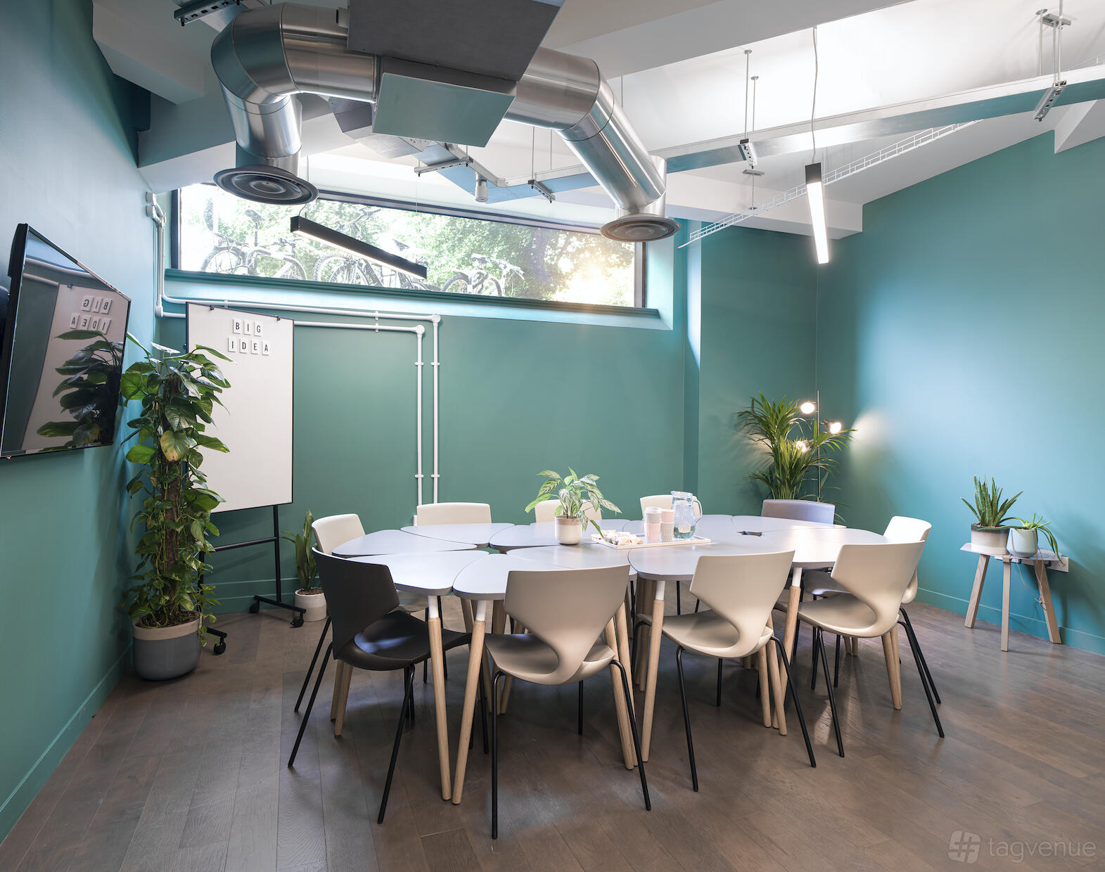 A meeting room with a round table, mixed chairs, large window, plants, and a wall-mounted screen at Uncommon Borough.