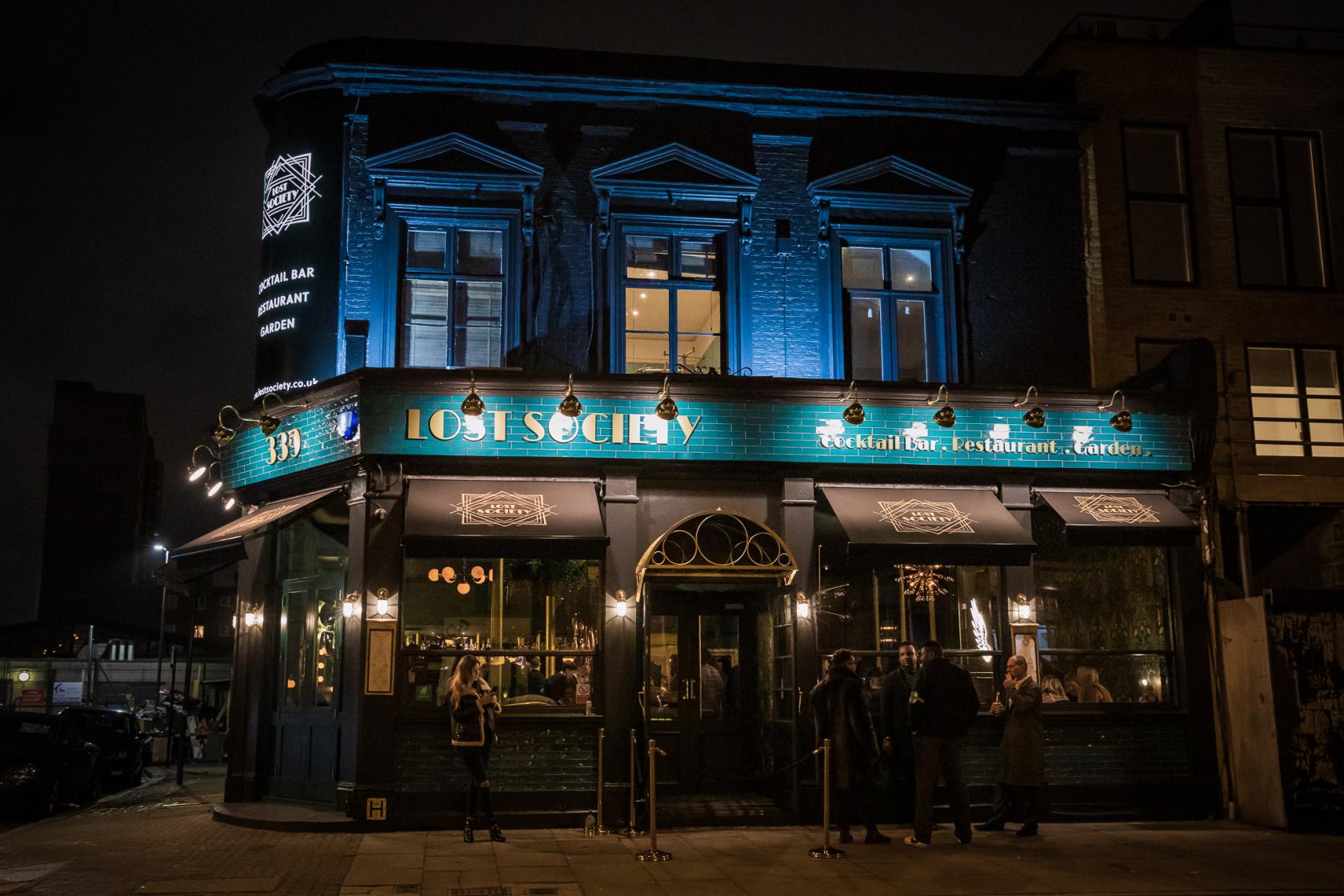 A pub and bar with illuminated signage, dark green facade, and large windows at Lost Society (Battersea SW11).