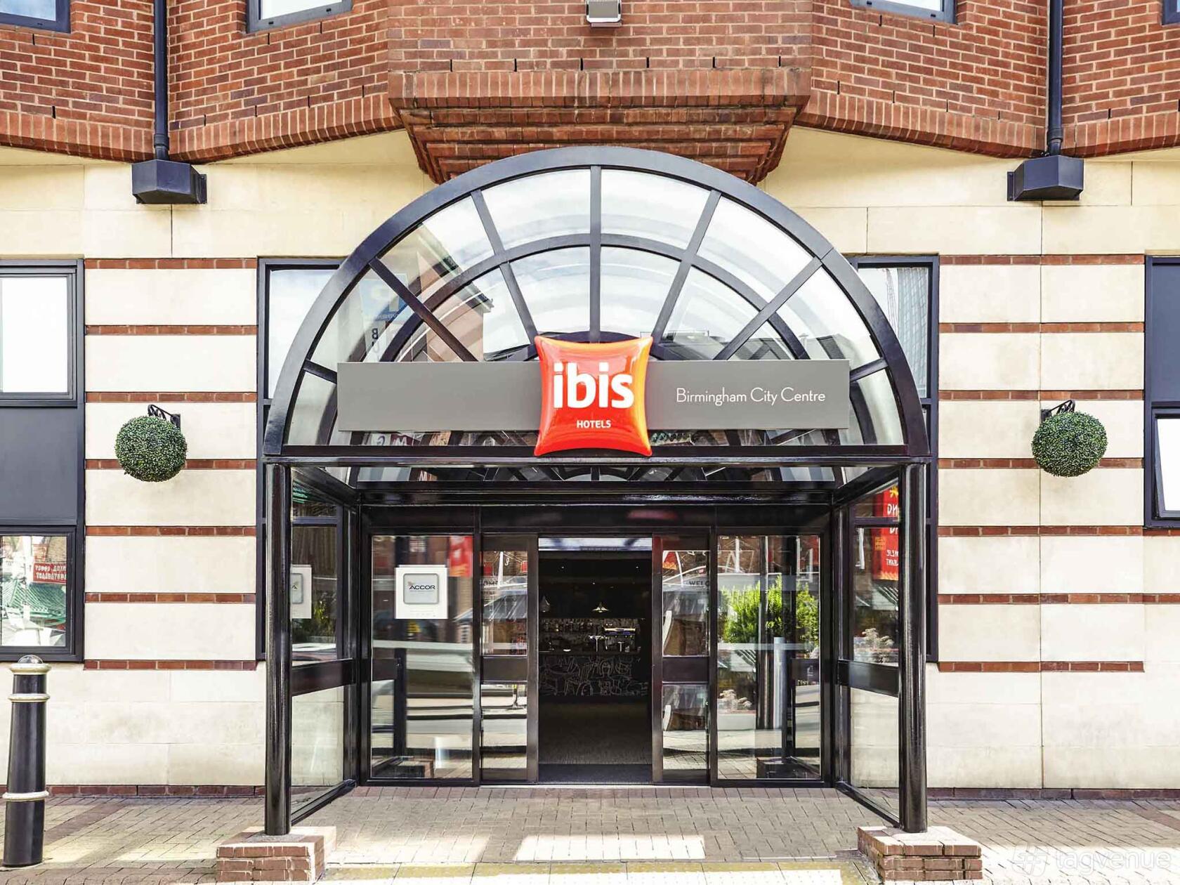 A hotel entrance with a curved glass awning, brick and stone facade, and glass doors at Ibis Birmingham New Street.