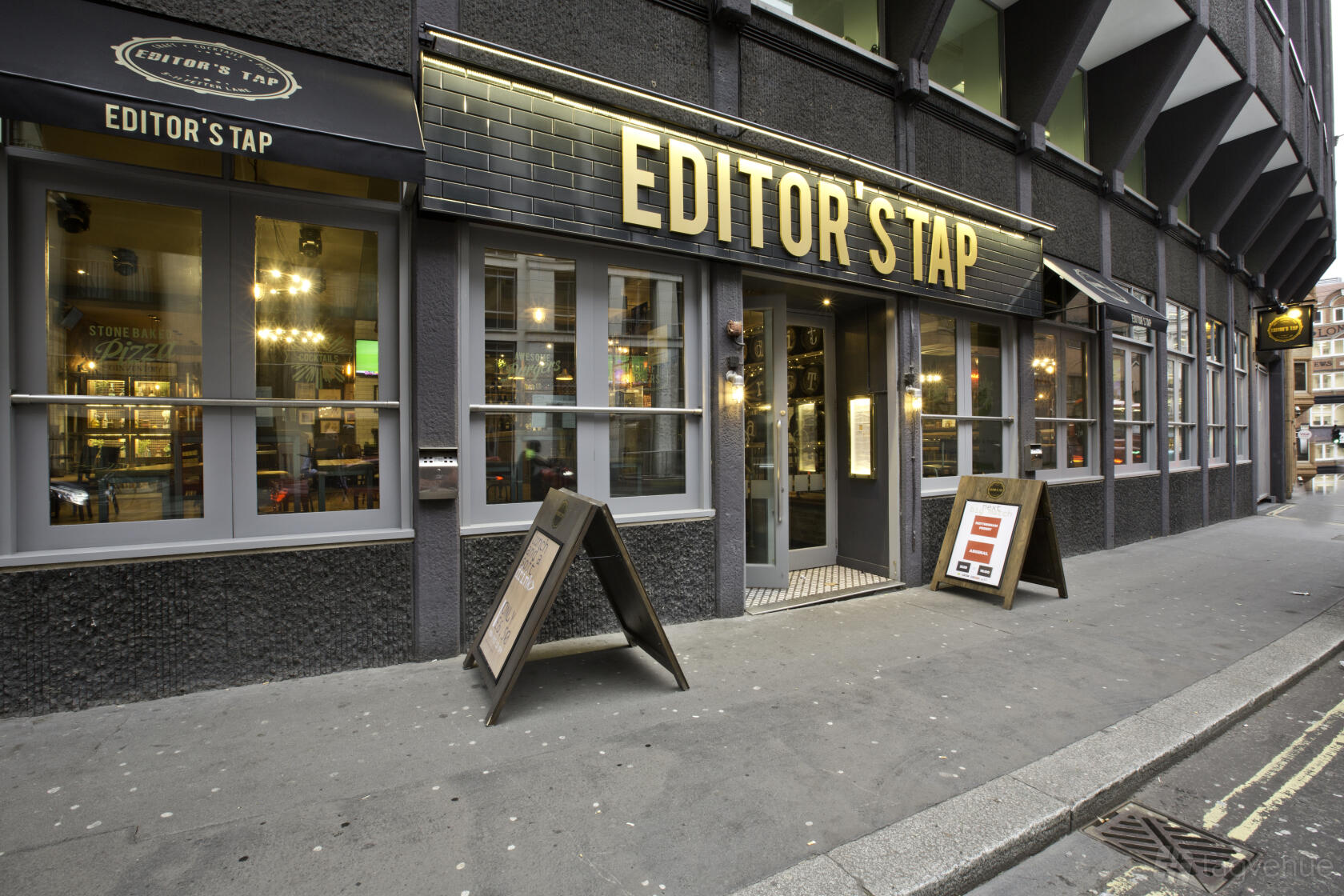 A pub with large front windows, illuminated signage, and sidewalk sandwich boards at Editors Tap.