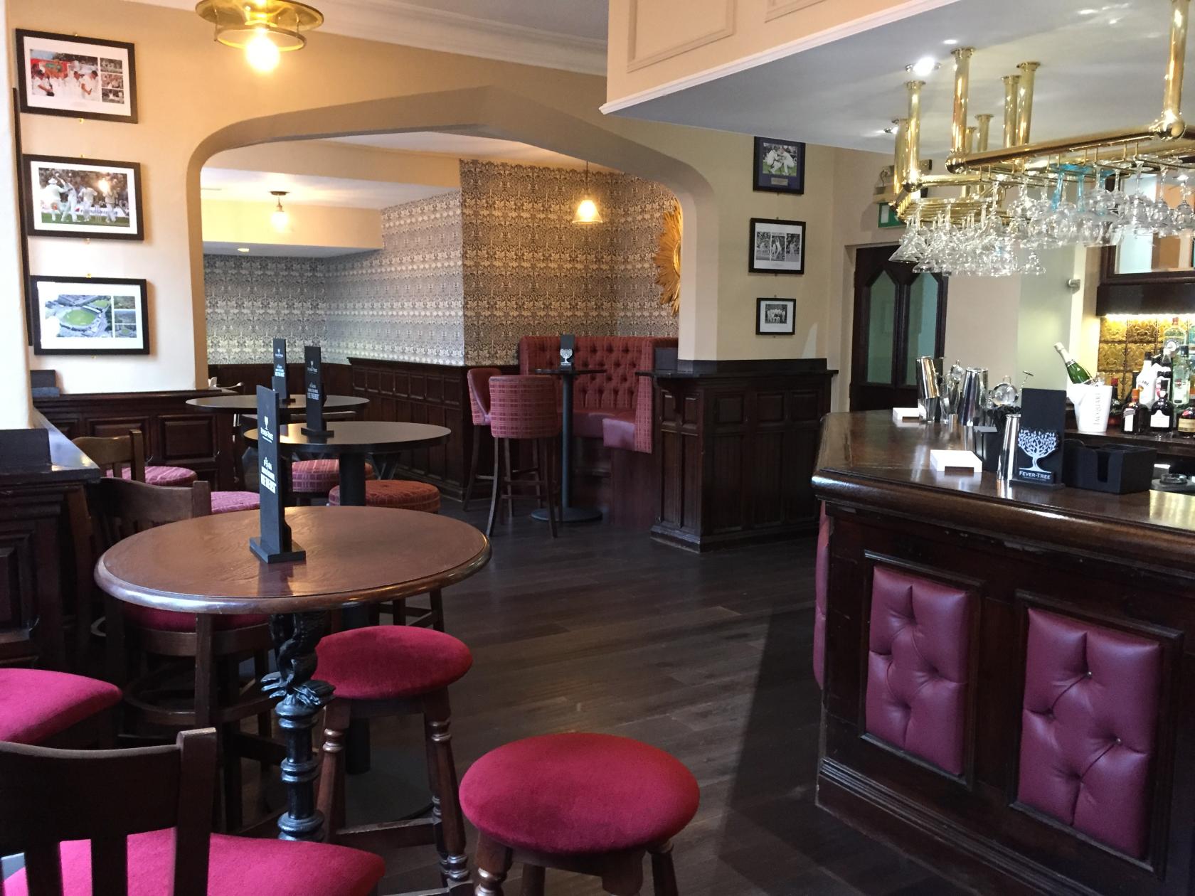 A hotel pub with dark wood paneling, red upholstered stools, round tables, and a glass rack above the bar at Plough & Harrow Hotel.