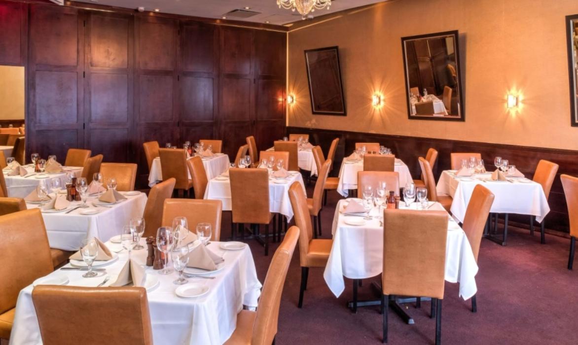 A dining room with tan leather chairs, white tablecloths, and wall sconces at Empire Steak House West.