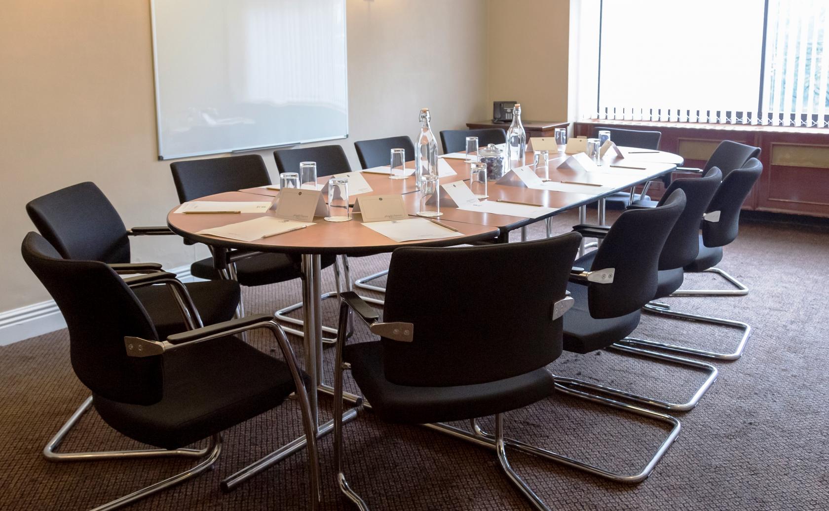 A meeting room with an oval conference table, black chairs, and a whiteboard at Plough & Harrow Hotel.