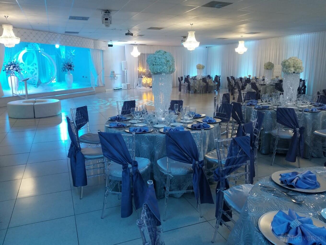 An event hall with round tables dressed in silver linens and blue napkins, chandeliers, and a draped stage at Royal Palace Banquet Hall.