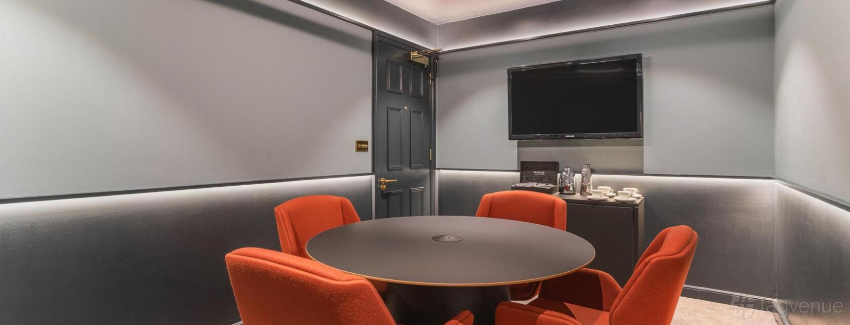 A meeting room with a round table, orange chairs, wall-mounted TV, and a tea and coffee station at 78-79 Pall Mall.