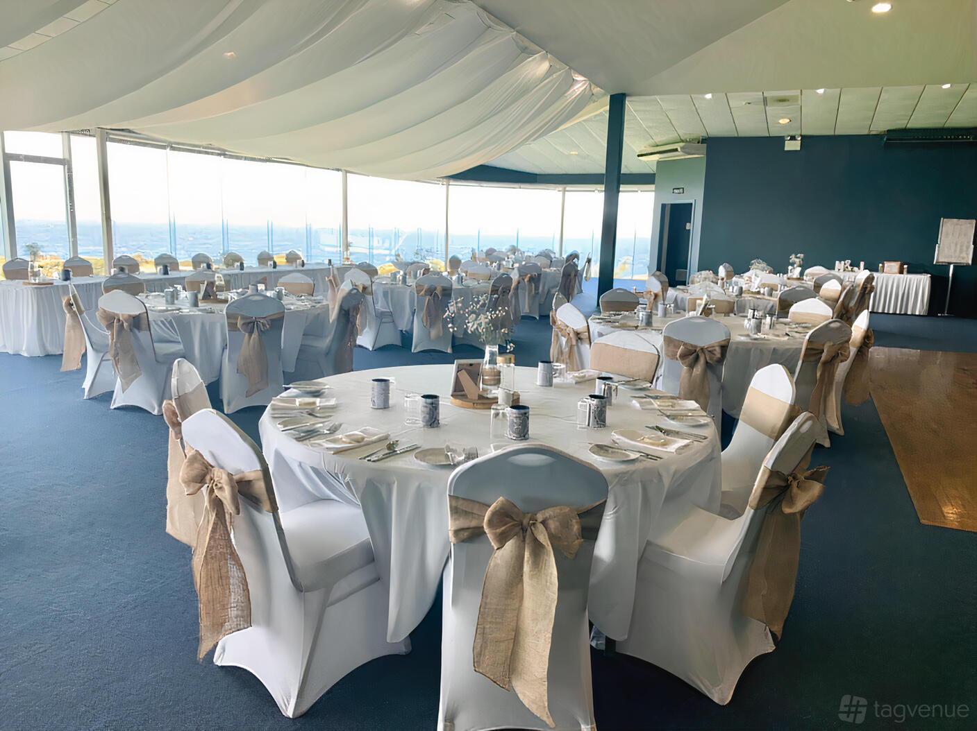 An event space with round tables draped in white linens and chair covers tied with burlap bows at Narooma Golf Club.
