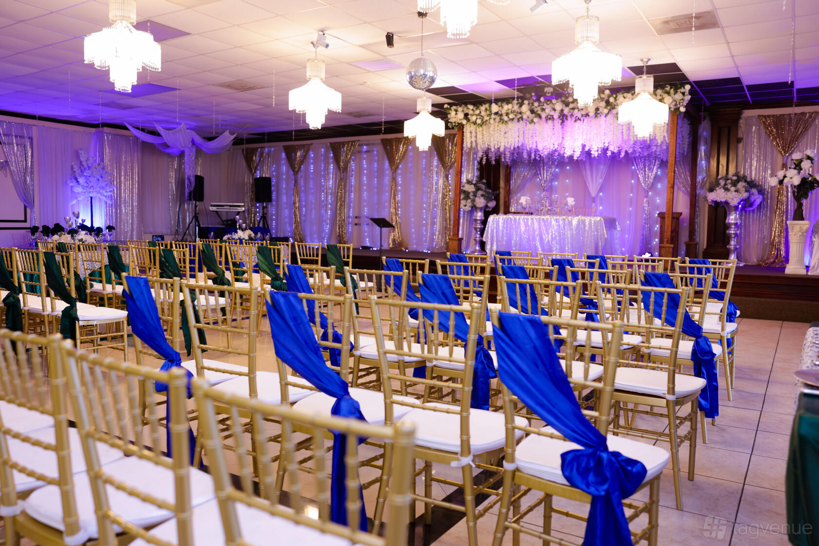 A decorated hall with gold chairs draped in blue sashes, chandeliers, and a floral stage at Lily Reception Hall.