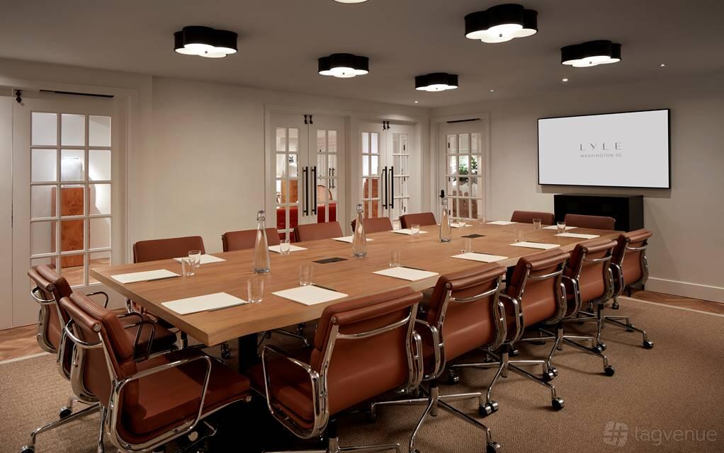 An event space in a hotel with a large wooden conference table, brown leather chairs, and a projector screen at Lyle Washington DC - Lower level.
