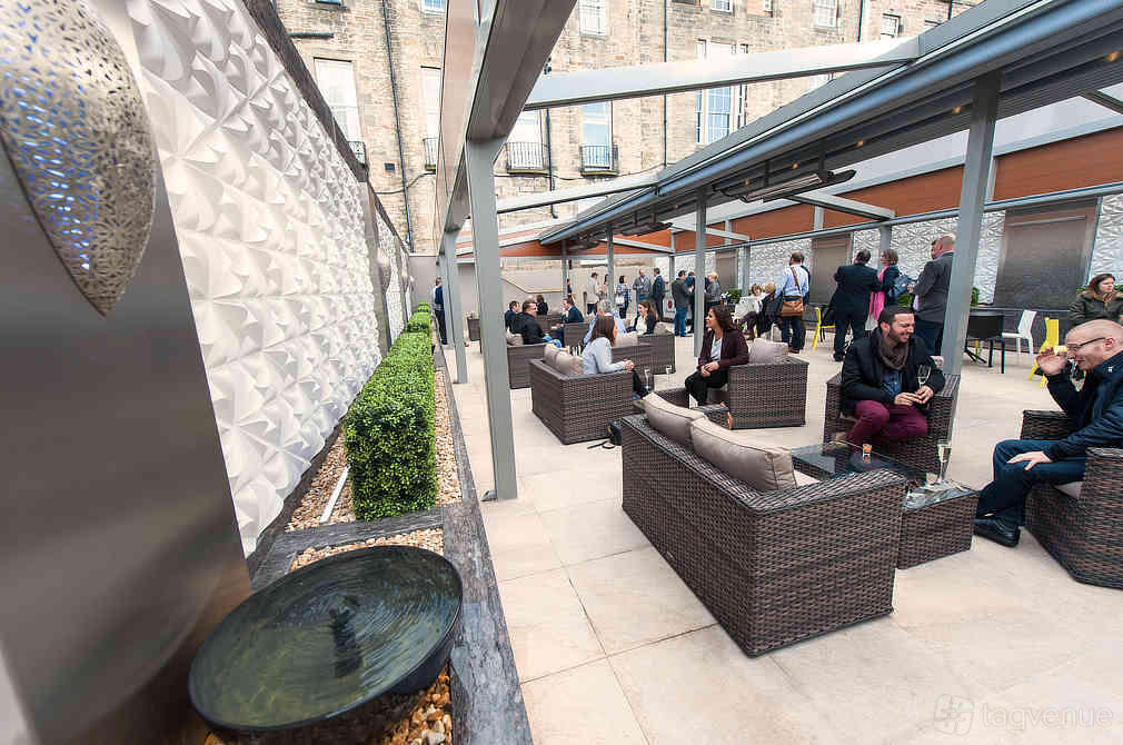 A hotel terrace with wicker lounge seating, covered pergolas, and a decorative water feature at The Place Hotel.