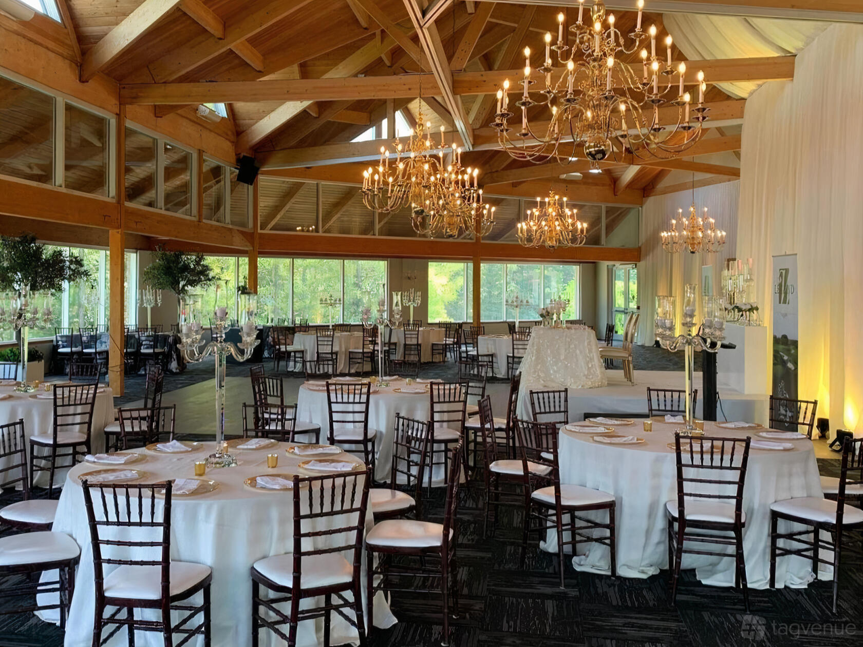 An event venue with floor-to-ceiling windows, exposed wooden beams, chandeliers, and round tables at Woodington Lake Golf Club.