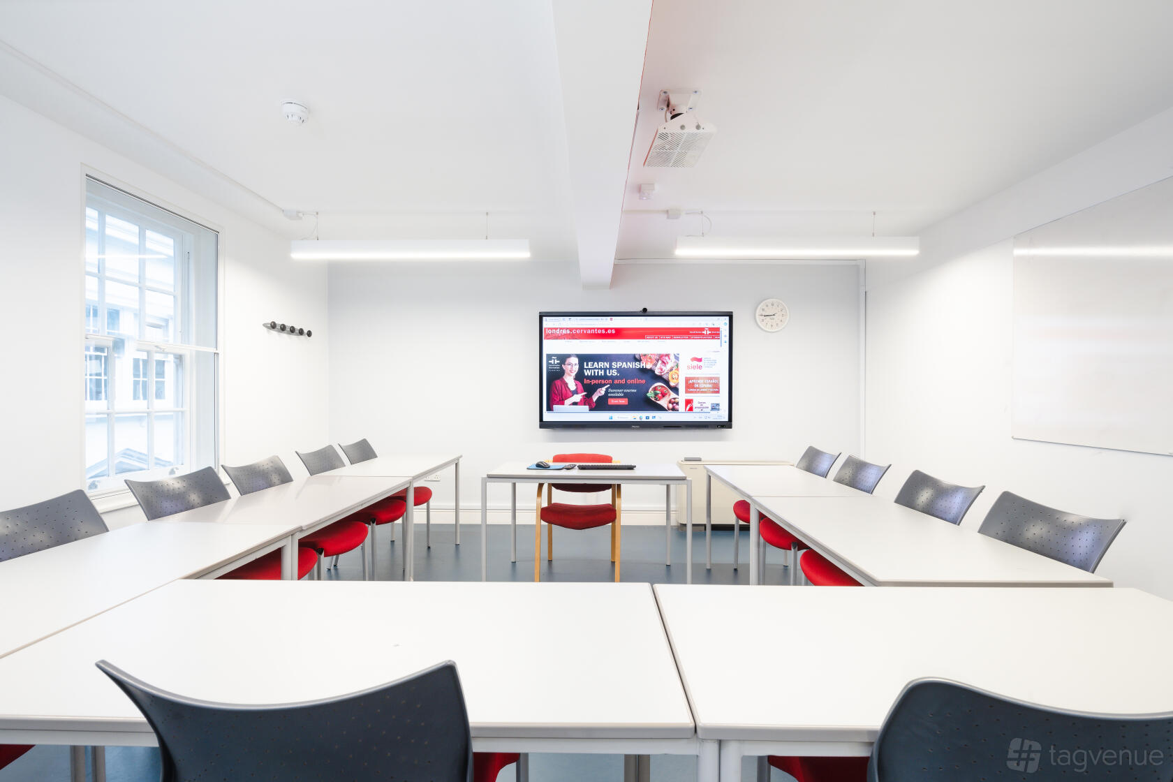 A conference classroom with U-shaped white tables, red and black chairs, large windows, and a digital screen at DEVEREUX COURT.