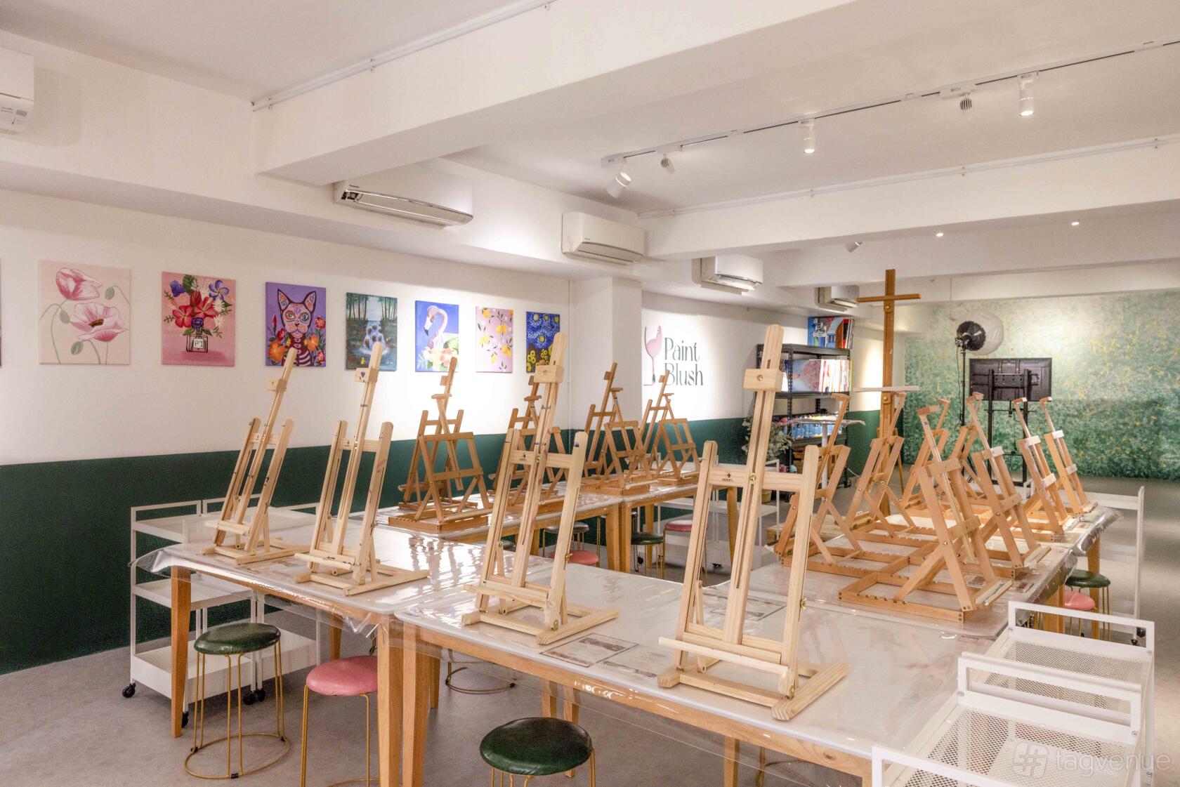 An art studio with easels on long tables, stools, colorful artwork on the walls, and bright lighting at PaintBlush.