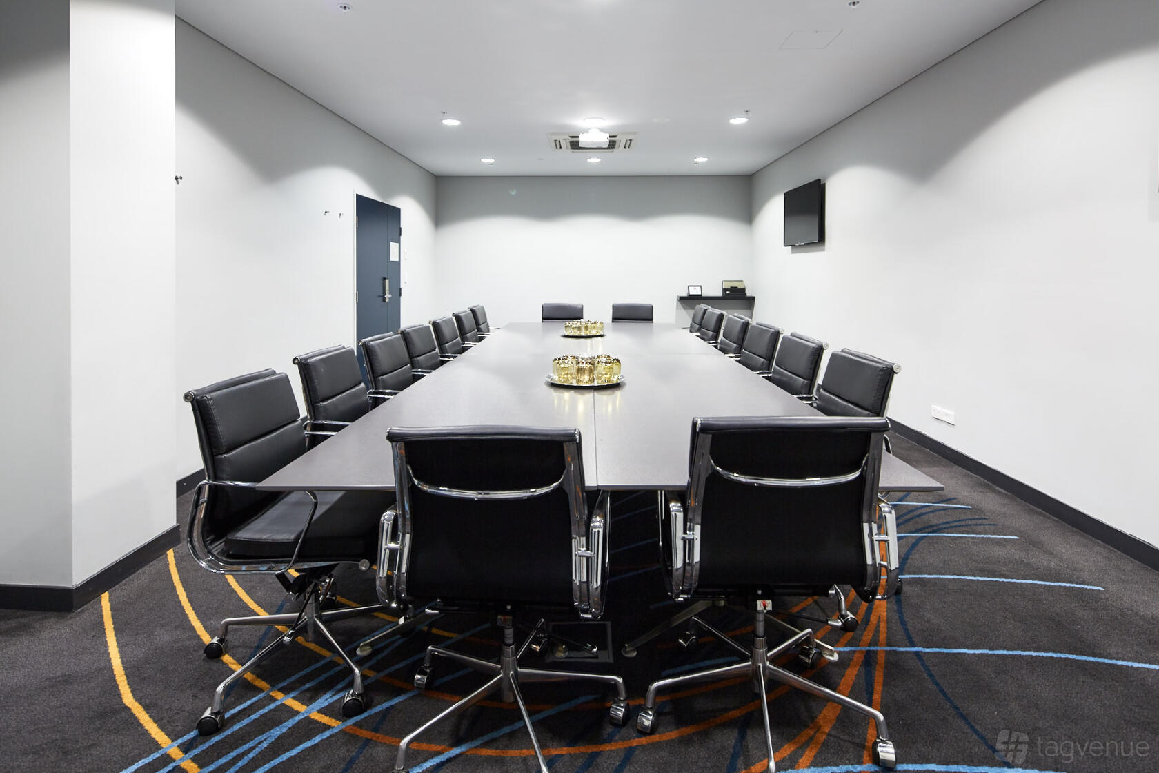 A boardroom with a long rectangular table, black leather chairs, and wall-mounted monitor at Rydges Sydney Airport Hotel.