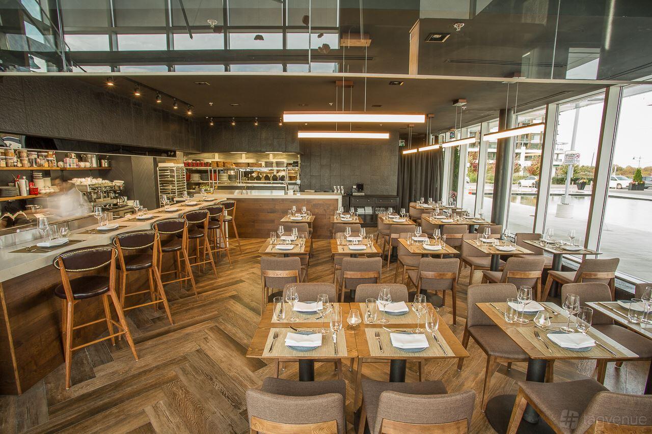 A restaurant dining area with wooden tables, upholstered chairs, an open kitchen, and large windows at Terre Rouge Craft Kitchen.