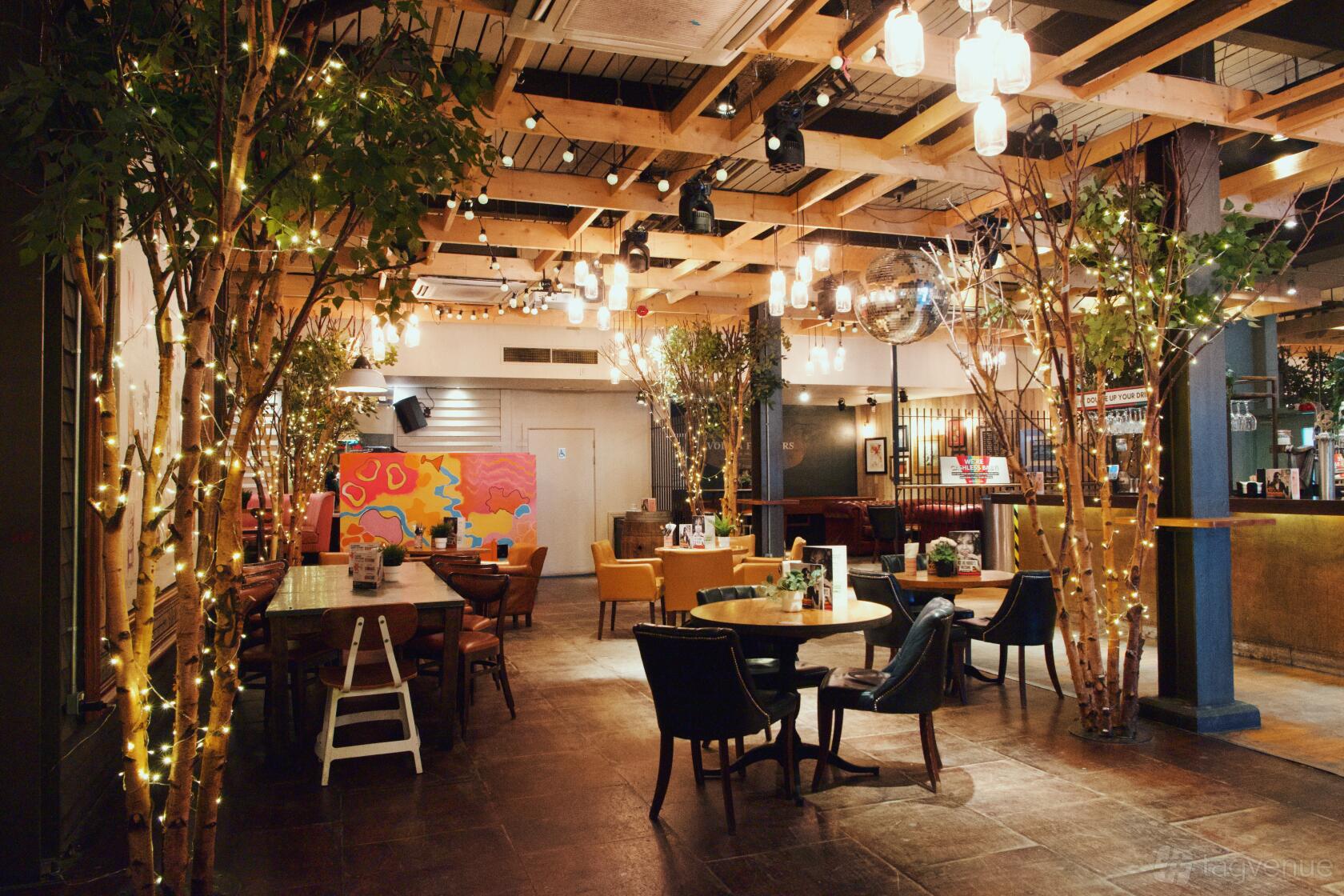 A bar with tree-like decor wrapped in string lights, exposed wooden beams, and eclectic seating at Revolution Cardiff.
