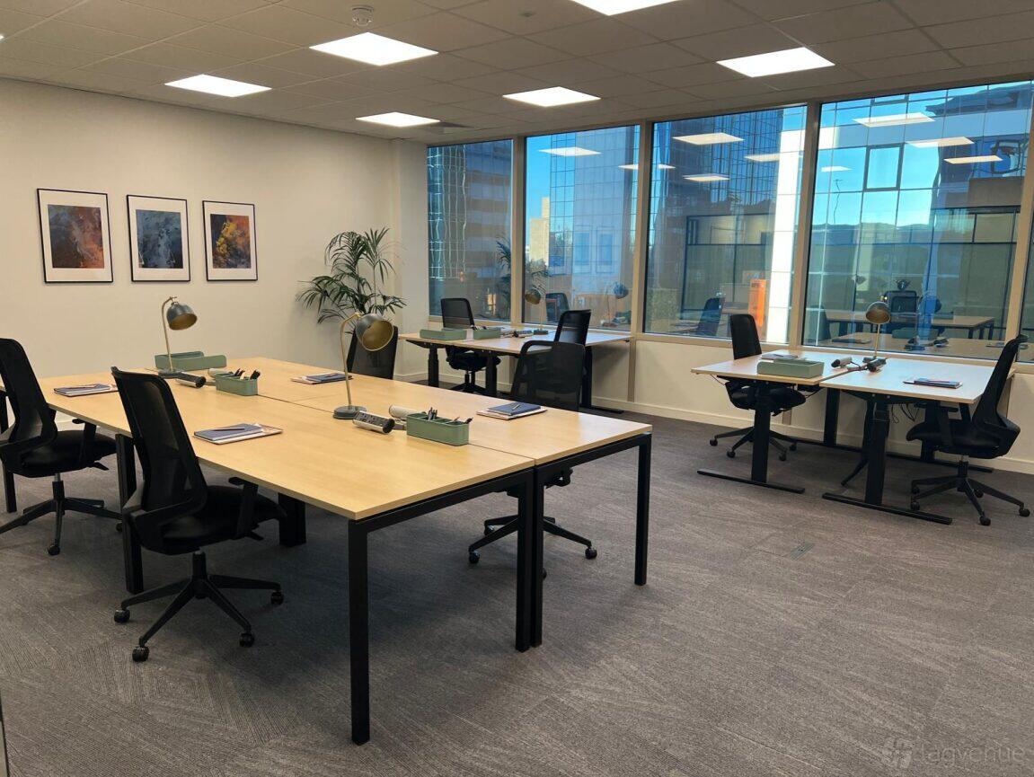 A boardroom with large windows, two long wooden tables, black chairs, and desk lamps at Serendipity Labs Manchester.