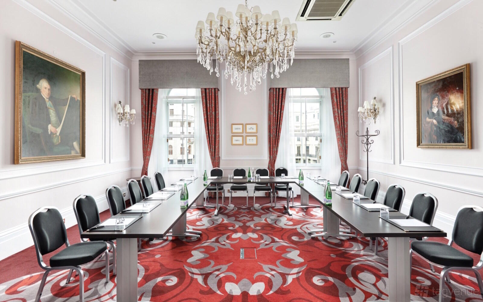 A hotel meeting room with U-shaped tables, ornate chandeliers, red patterned carpet, and large windows at Clermont Charing Cross.