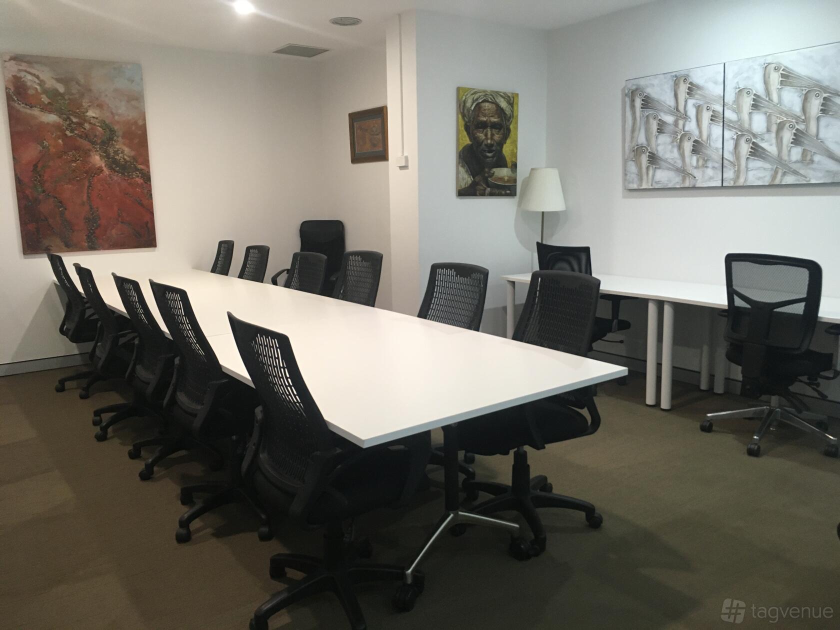 A training room with long white tables, black mesh chairs, carpeted flooring, and wall art at A23.