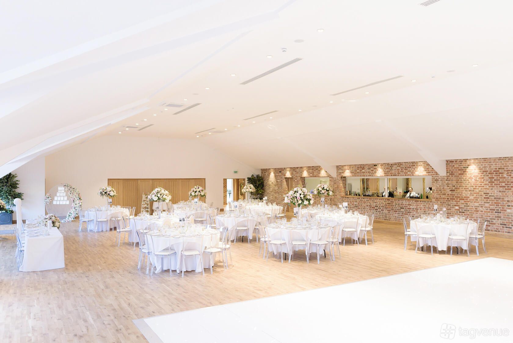 A banqueting hall with round tables draped in white linens, floral centerpieces, and exposed brick accents at Hatfield Place.
