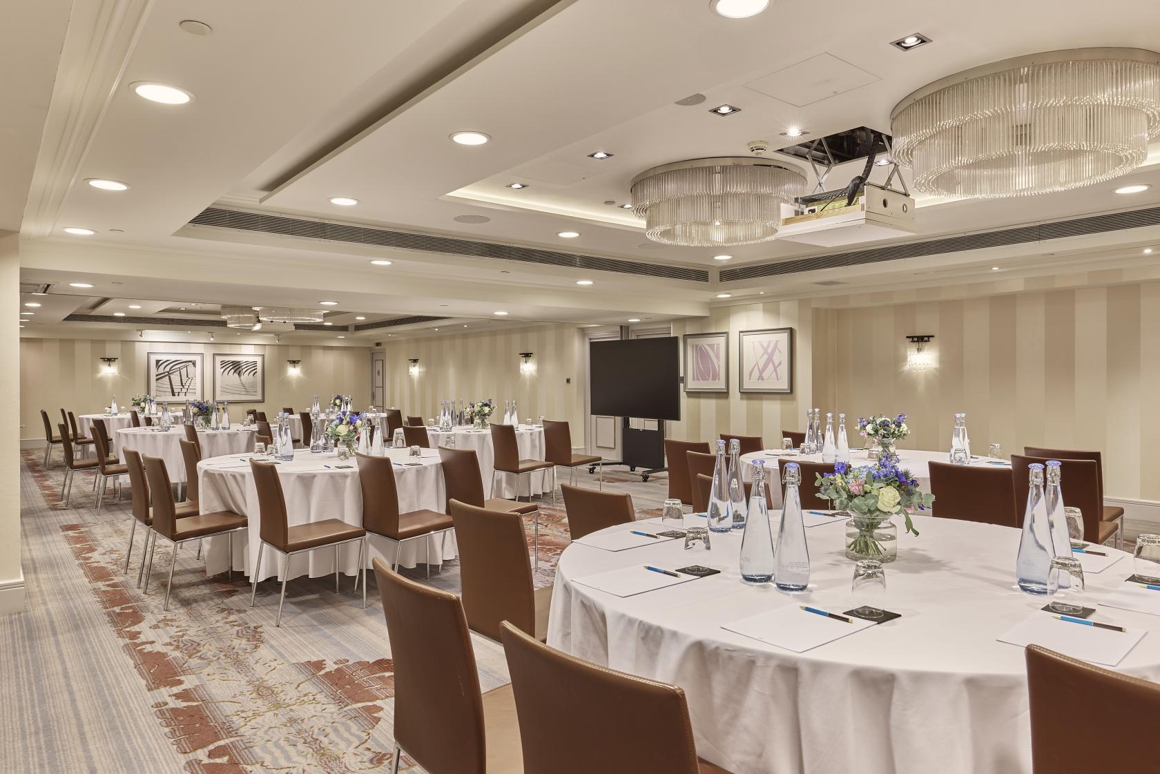 An event space in a hotel with round tables, brown chairs, neutral striped walls, and ceiling chandeliers at Meetings & Events Venues.