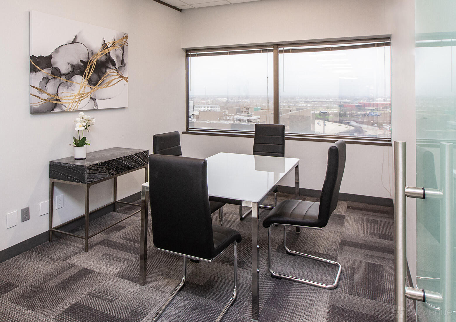 A meeting room with a glass table, four black chairs, large window, and abstract artwork at Lyric Works.