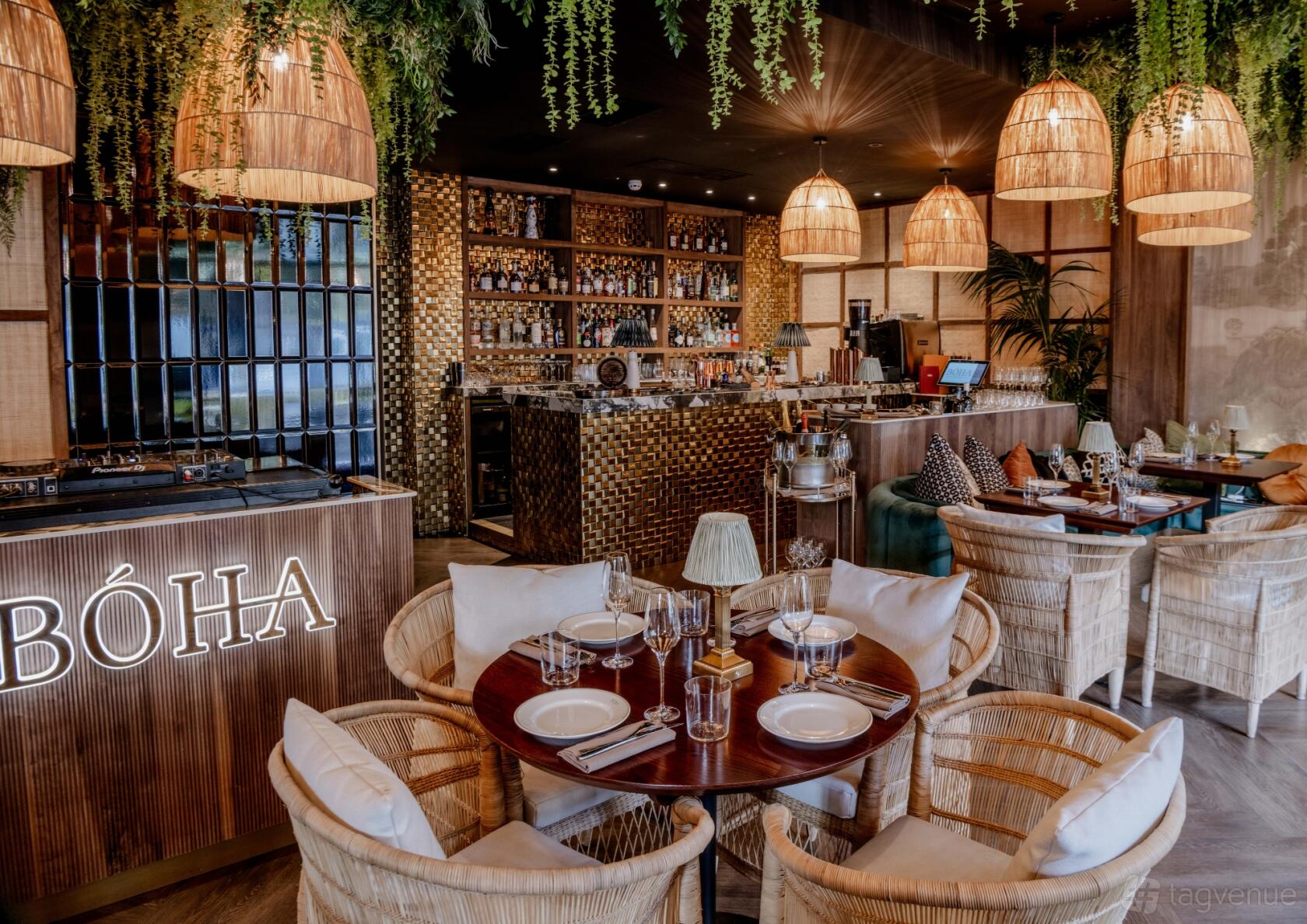 A bar in a restaurant with wicker pendant lights, greenery overhead, and gold mosaic tile behind the bar at BÓHA London.