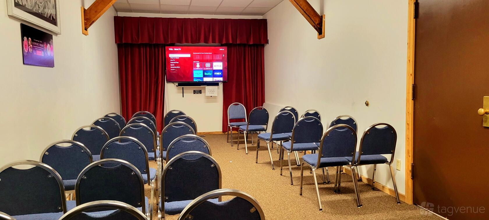An event space with blue cushioned chairs, a red curtain backdrop, and a wall-mounted TV at Western Museum of Mining and Industry.