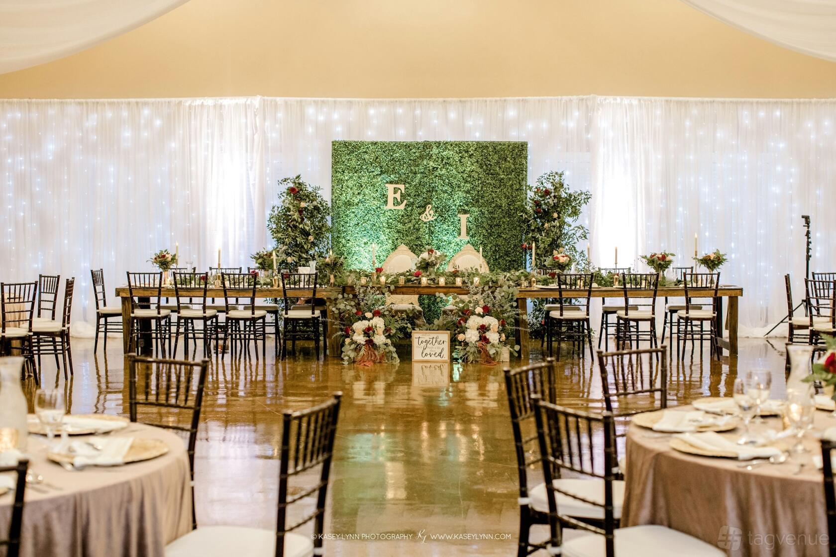An event venue with a greenery wall backdrop, string lights, and round tables with beige linens at Oak Tree Manor.