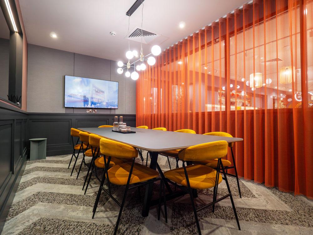 A meeting room with a long table, yellow chairs, modern pendant lights, and an orange sheer divider at Colony Fabrica.