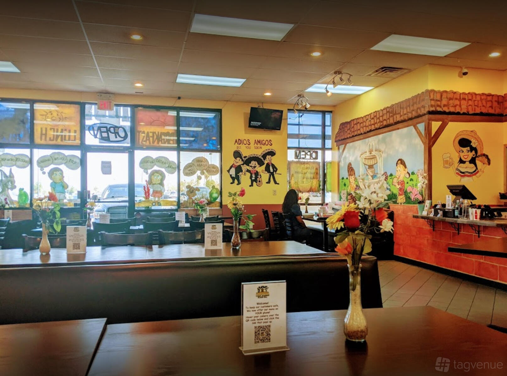 A restaurant with colorful murals, large front windows, and wooden tables with small flower arrangements at Tres Amigos Mexican Grill.