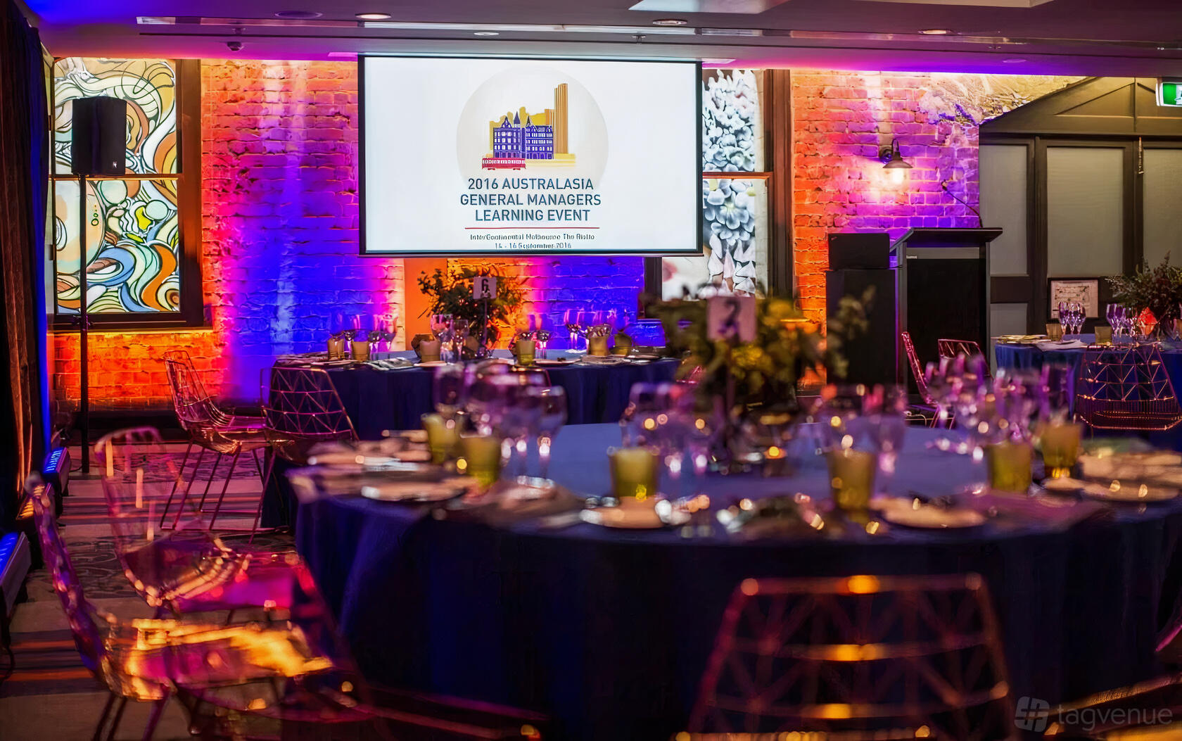 An event room with exposed brick walls, colorful uplighting, round tables with blue linens, and acrylic chairs at InterContinental Melbourne.