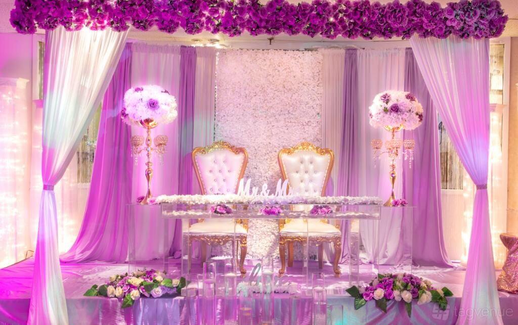 A ballroom with a floral backdrop, gold-trimmed chairs, purple draping, and floral arrangements at Charisma Ballroom.