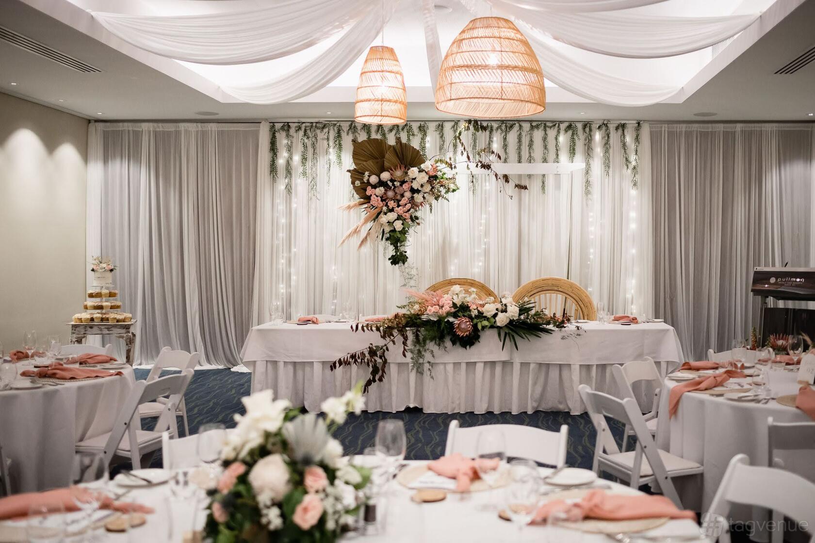 A hotel ballroom with draped white ceilings, floral arrangements, and round tables set for an event at Pullman Bunker Bay.
