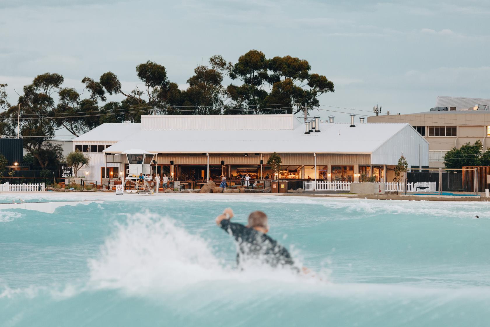 A restaurant with a white facade, large windows, and outdoor seating, located beside an artificial surfing lagoon at Three Blue Ducks Melbourne.
