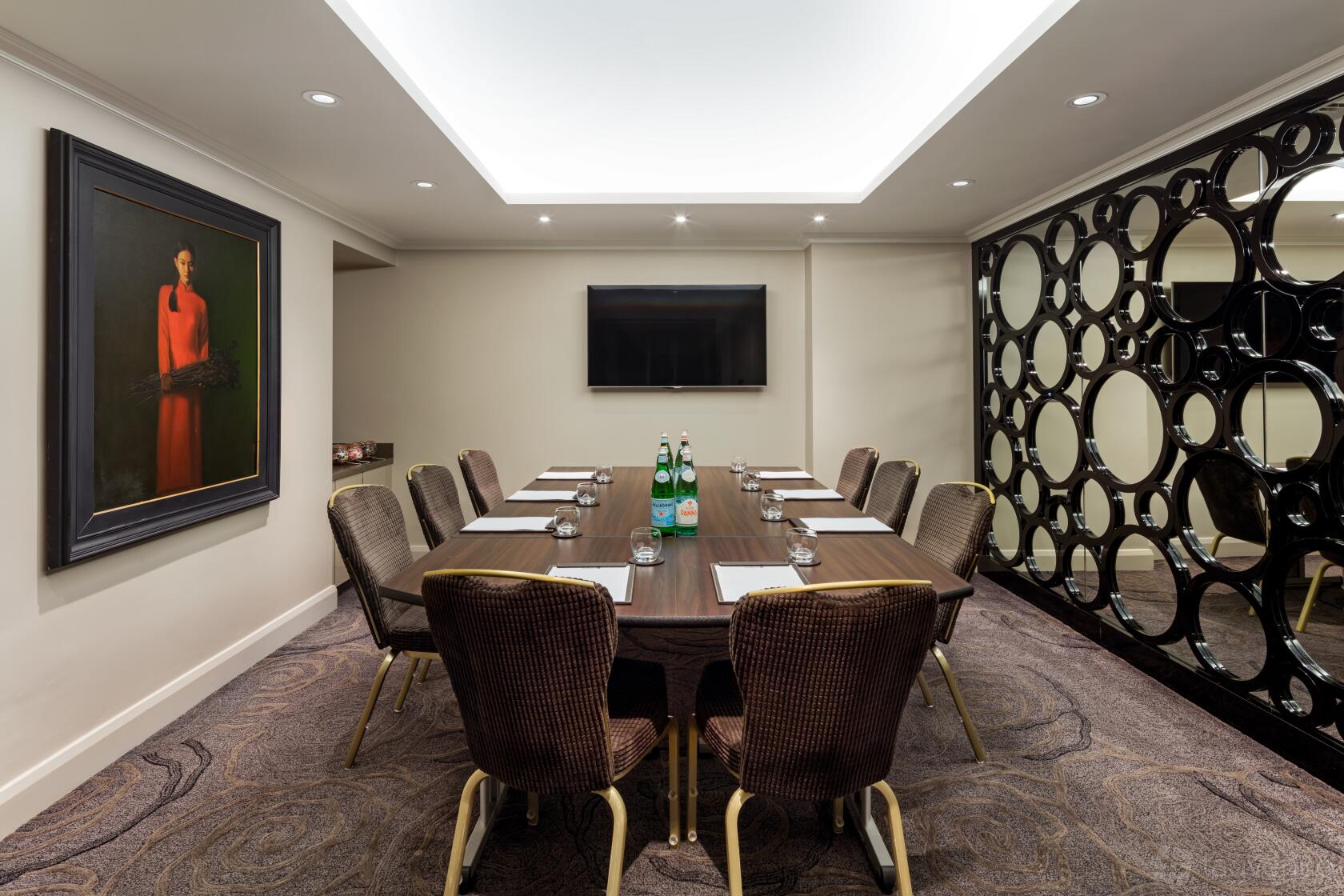 A meeting room with a rectangular table, cushioned chairs, wall-mounted TV, and decorative mirror panels at Radisson Blu Hotel, London South Kensington.