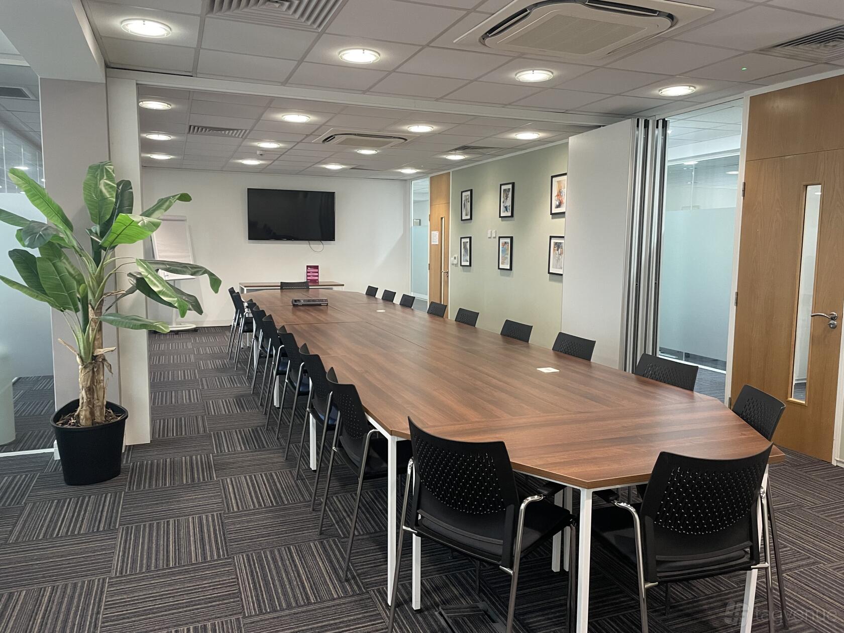 A boardroom with a long wooden conference table, black chairs, wall-mounted TV, and potted plant at Citibase Local Birmingham Business Park.