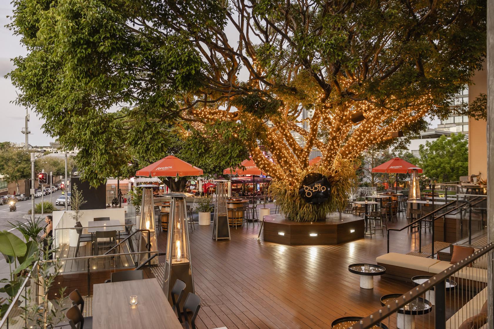 A restaurant terrace with a large fig tree wrapped in string lights, wooden decking, and outdoor seating at Bar Gusto.