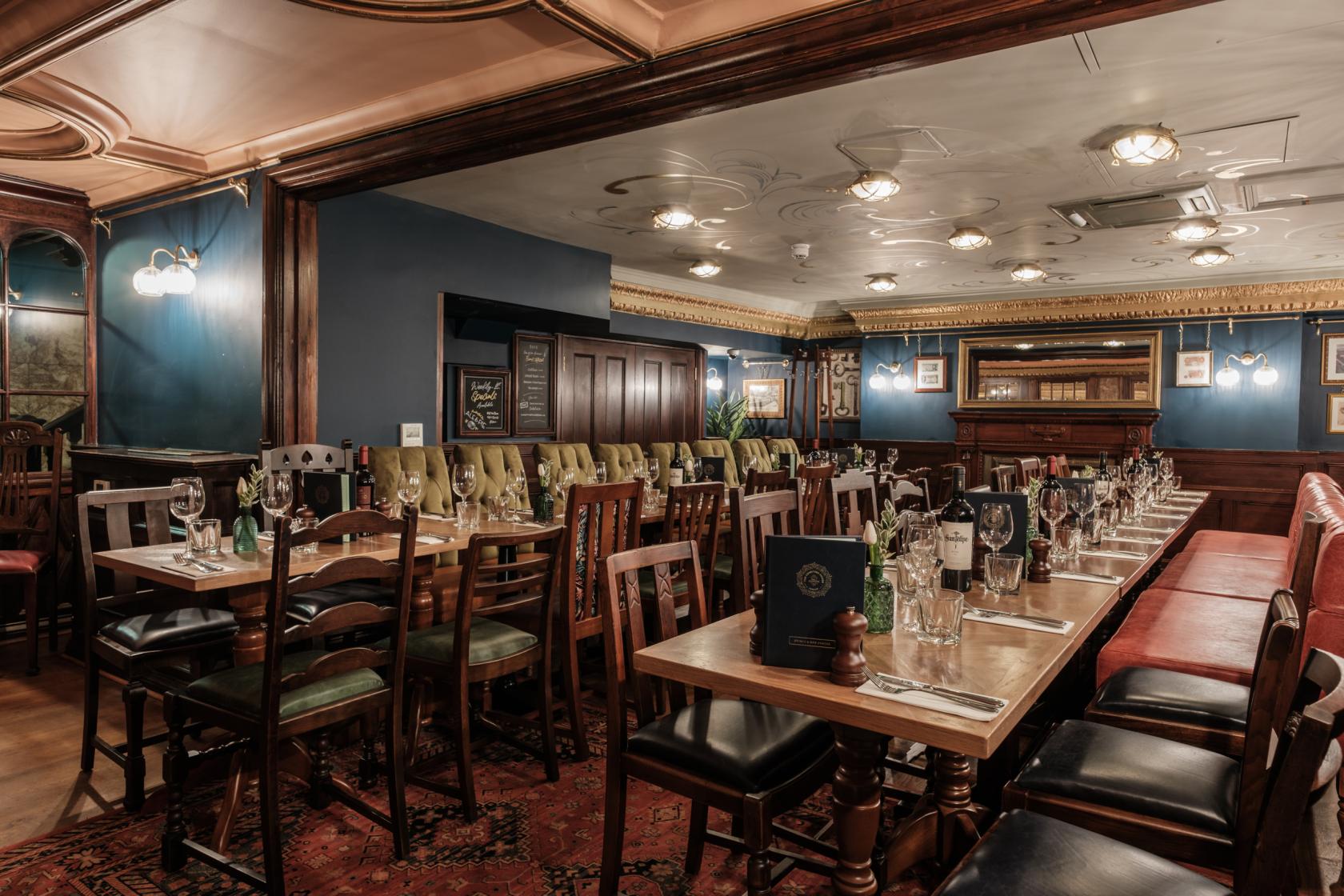 A restaurant with tufted leather banquettes, wooden tables set for dining, and ornate ceiling at The Counting House.