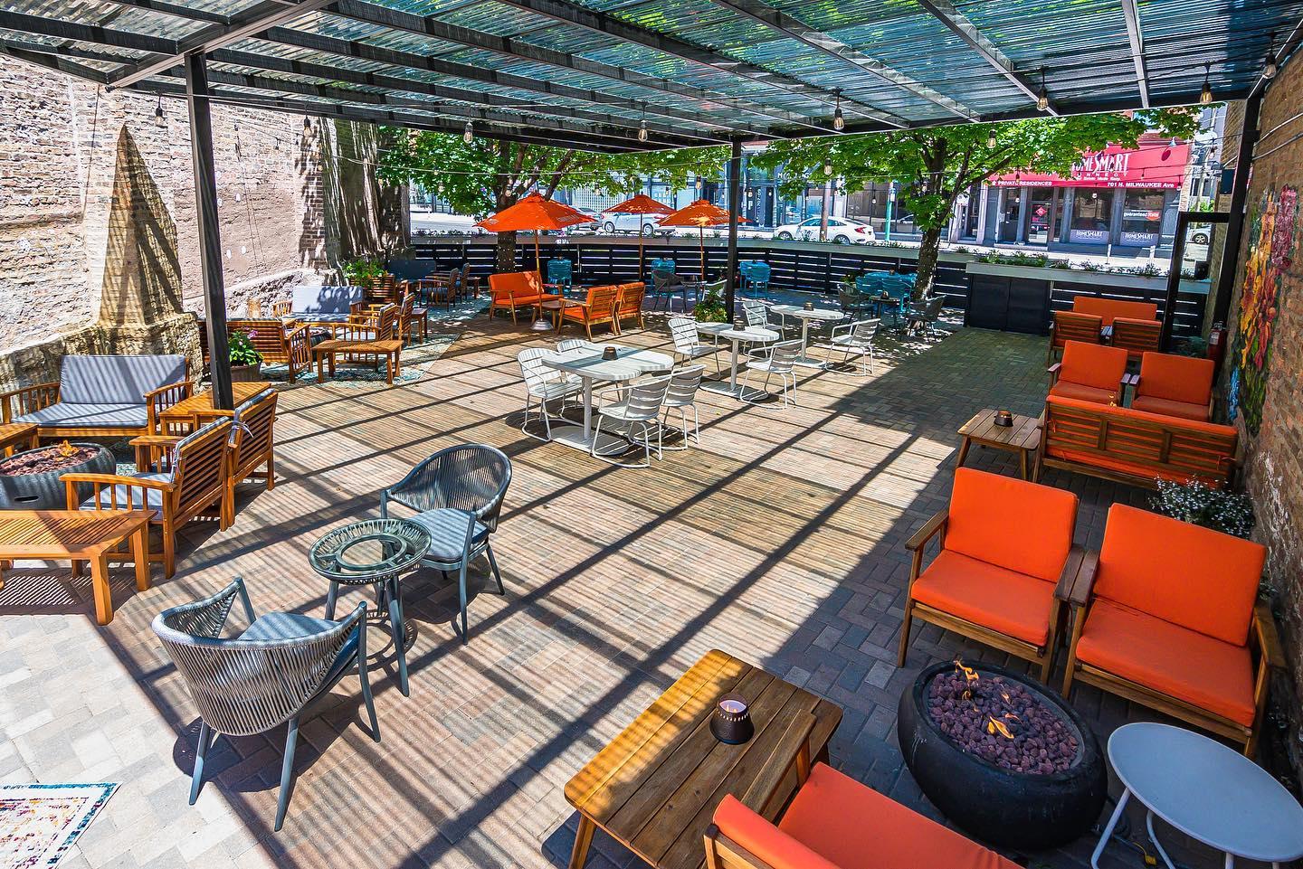 An outdoor bar patio with orange cushioned seating, umbrella-covered tables, and a partially shaded deck at The Dandy Crown.