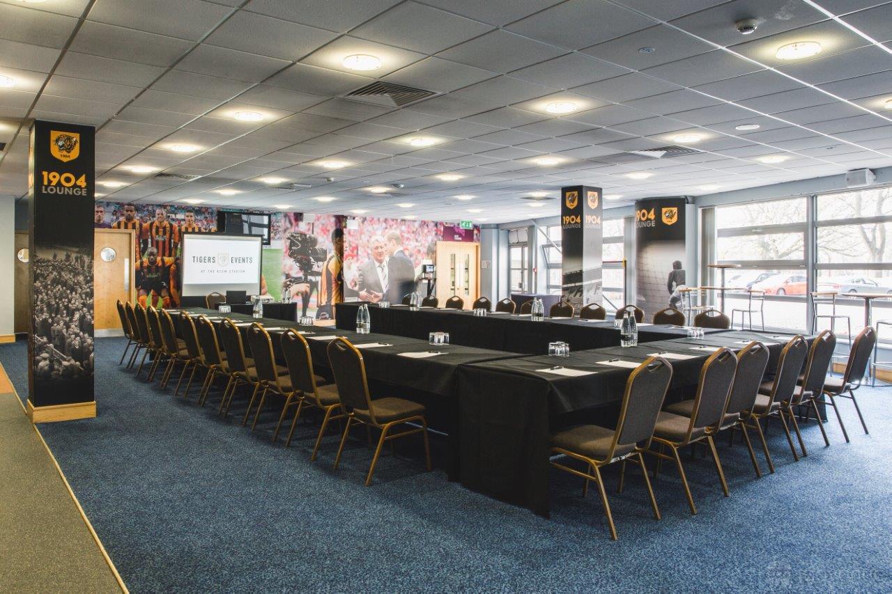 A lounge with U-shaped tables, padded chairs, carpeted flooring, and sports murals at MKM Stadium, Hull.