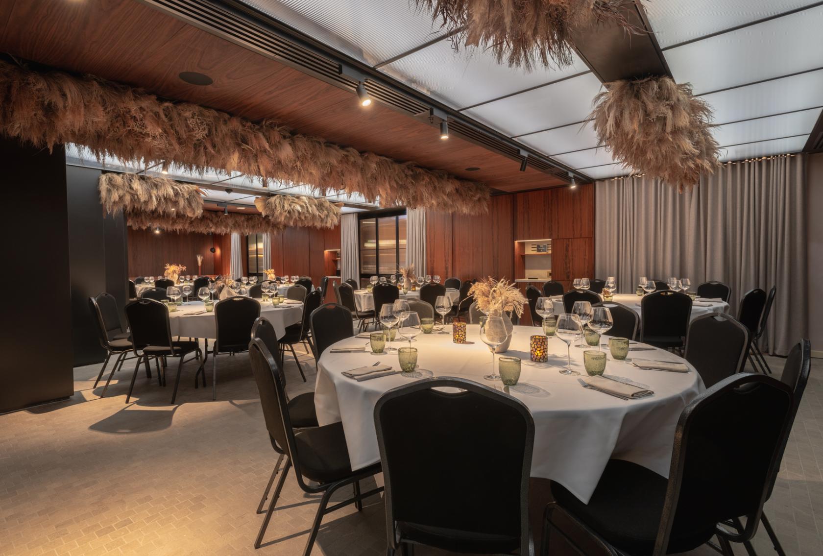 A hotel event room with round tables set for dining, pampas grass ceiling décor, and wood-paneled walls at Canopy by Hilton London City.