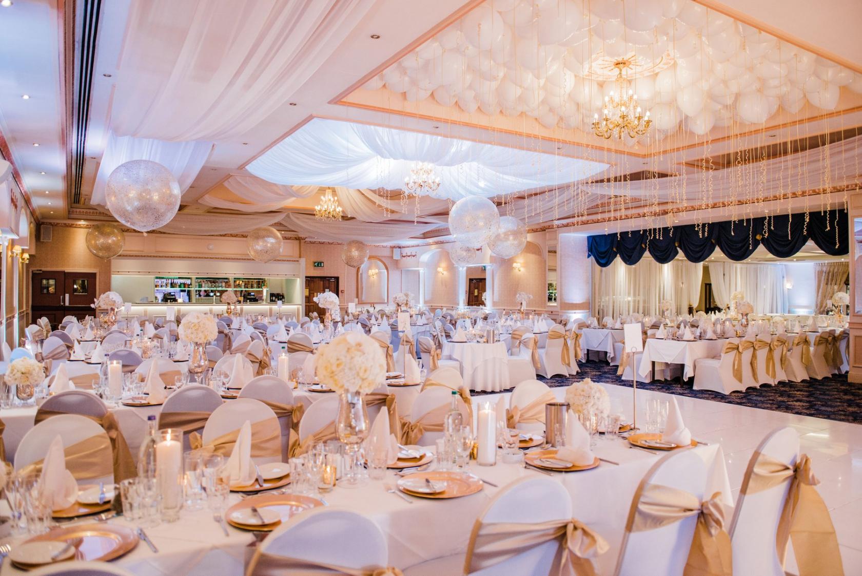 A hotel ballroom with draped white ceilings, chandeliers, round tables set with gold accents at New Continental Hotel.