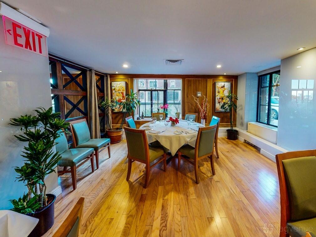 A dining room with round tables, green upholstered chairs, wood floors, and large windows at Tudor City Steakhouse.