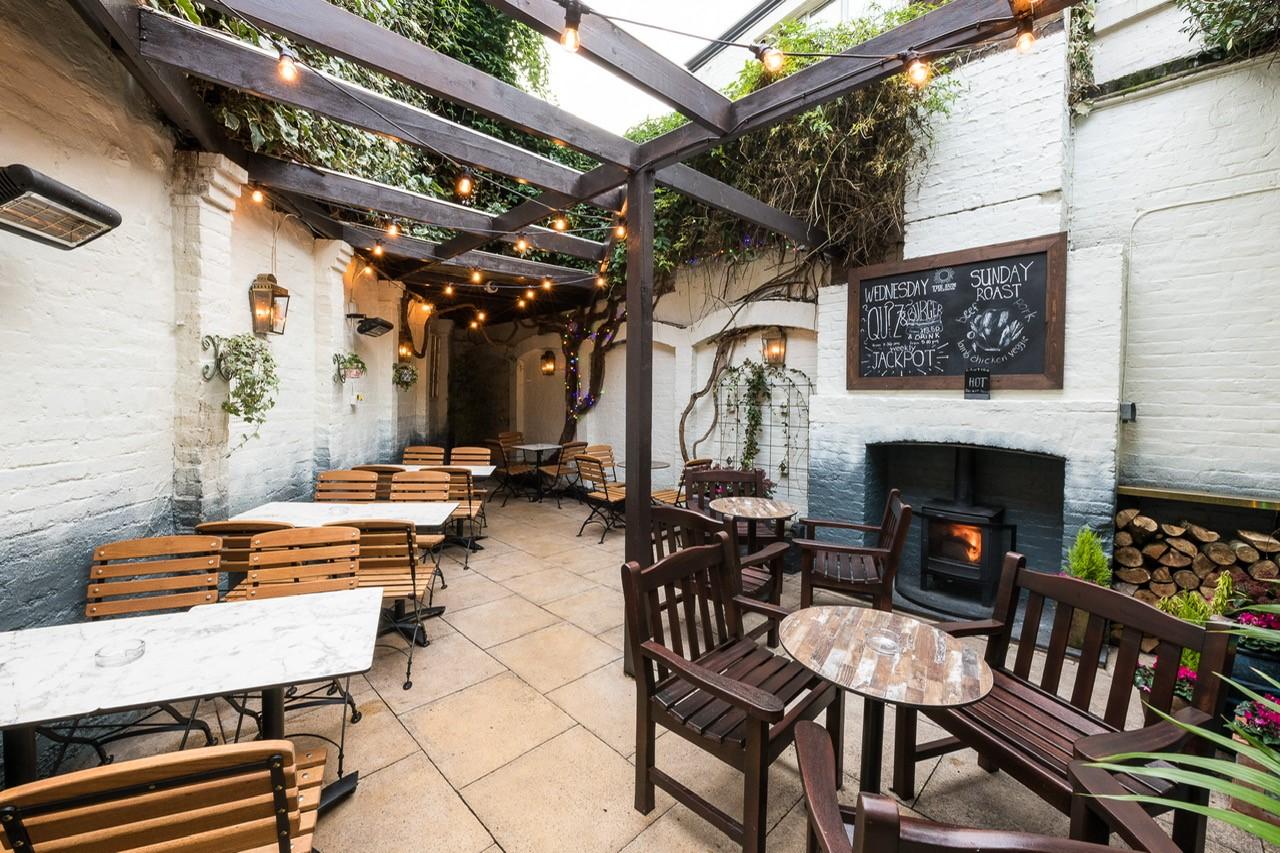 A pub terrace with wooden tables and chairs, string lights, a fireplace, and chalkboard menus at The Sun In Splendour.