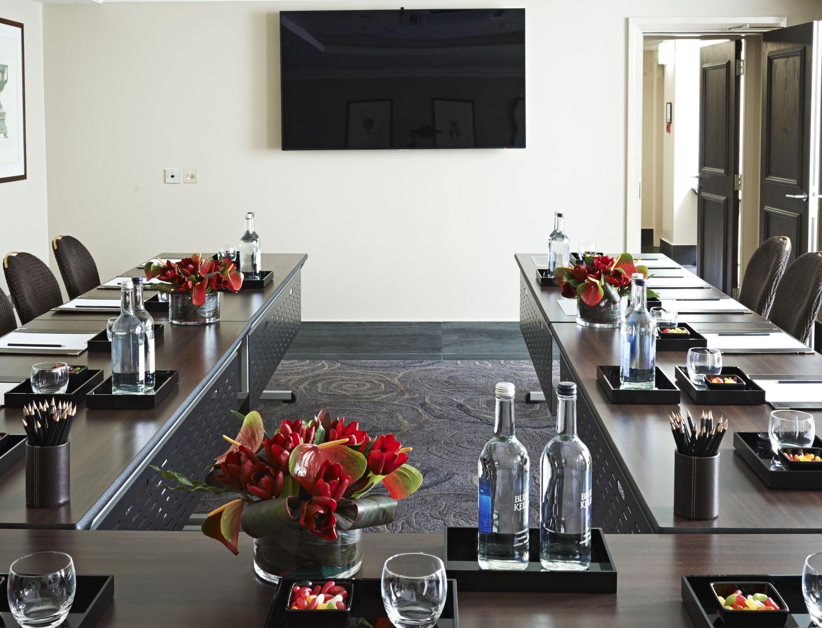 A hotel meeting room with U-shaped tables, bottled water, red floral centerpieces, and notepads at Radisson Blu Hotel, London South Kensington.