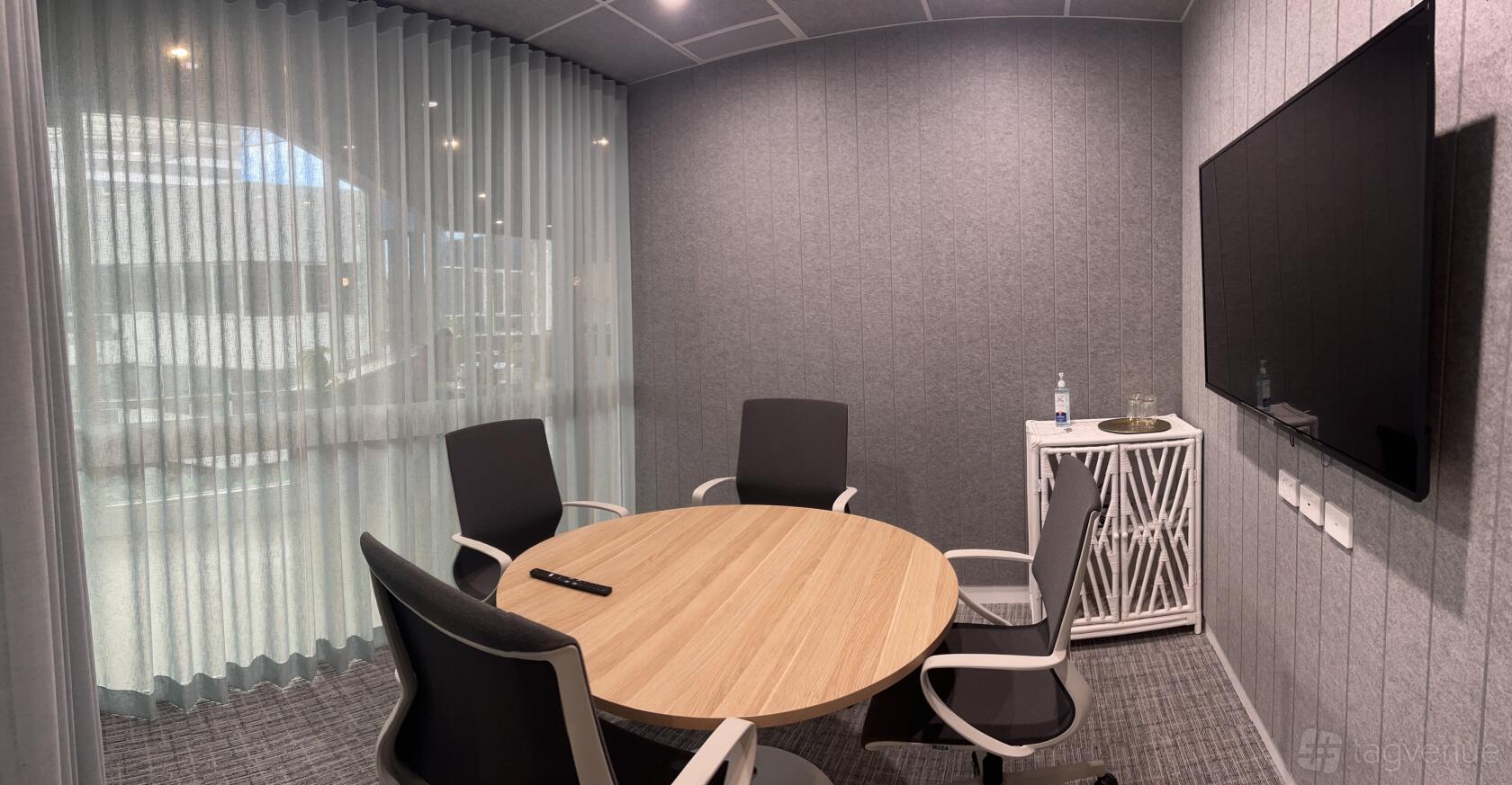 A meeting room with a round wooden table, four chairs, grey acoustic walls, and sheer curtains at @Workspaces - Milton.