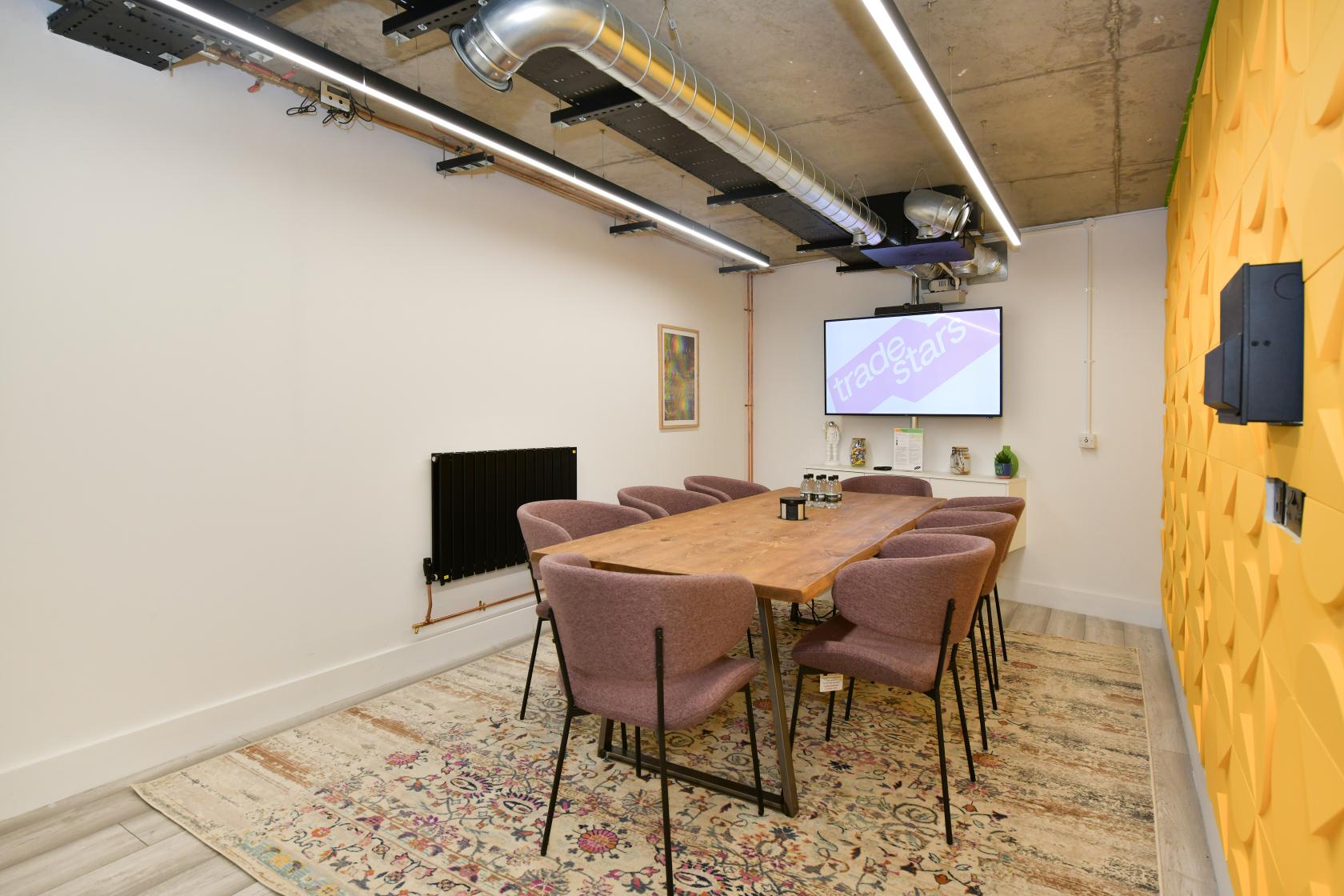 A meeting room with a long wooden table, eight upholstered chairs, yellow textured wall, and a large screen at Tradestars Hackney Wick - Bookable Spaces.