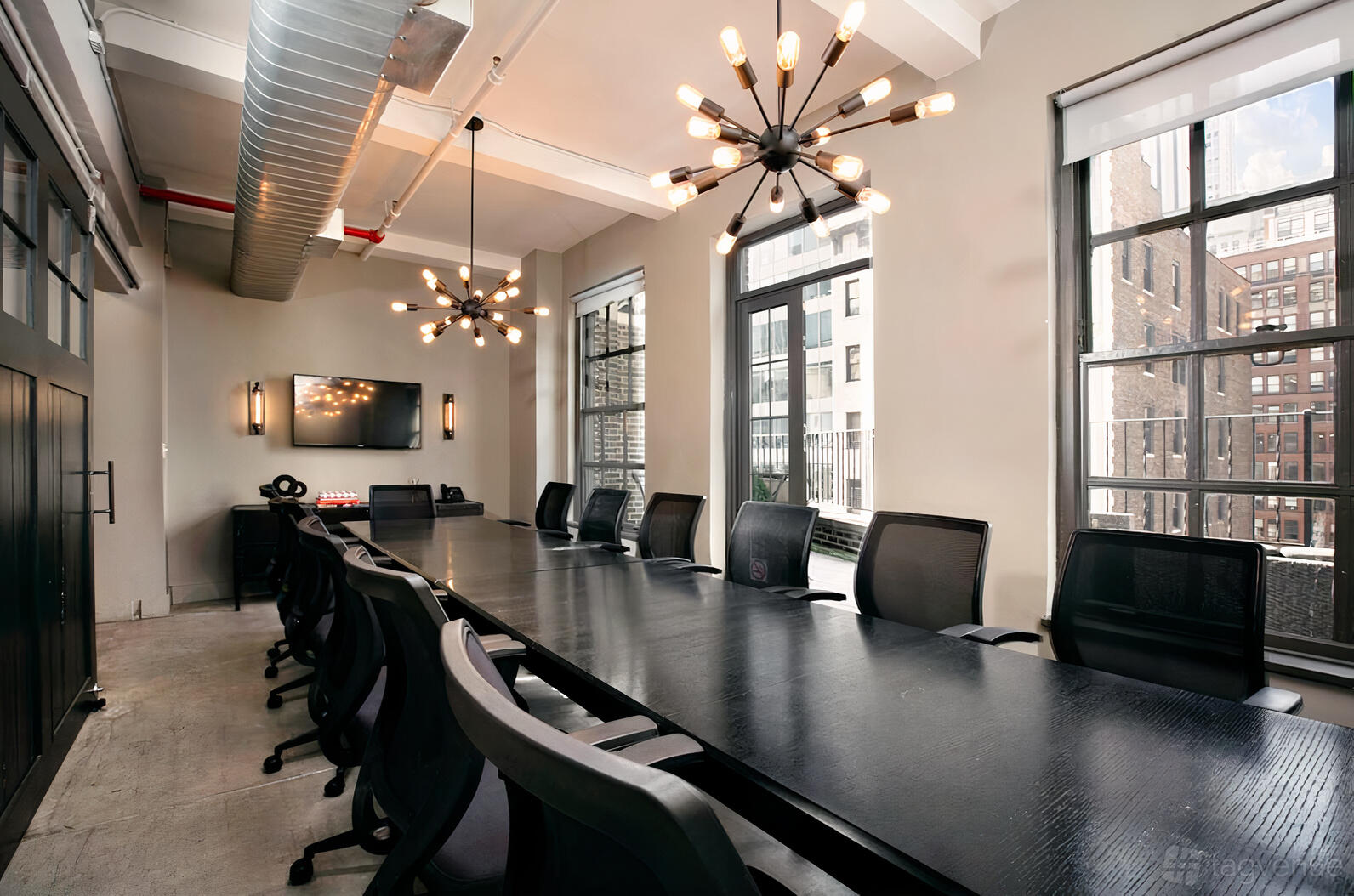 A meeting room with a long black conference table, modern chandeliers, and floor-to-ceiling windows at WorkHouse NYC - Midtown.