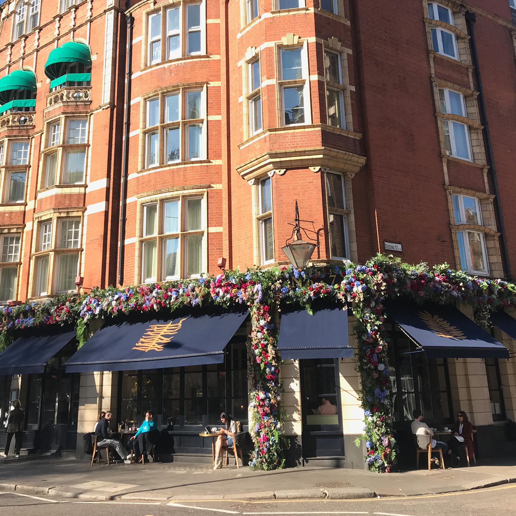 A cocktail bar exterior with red brick facade, large windows, blue awnings, and floral garlands at The Botanist Sloane Square.