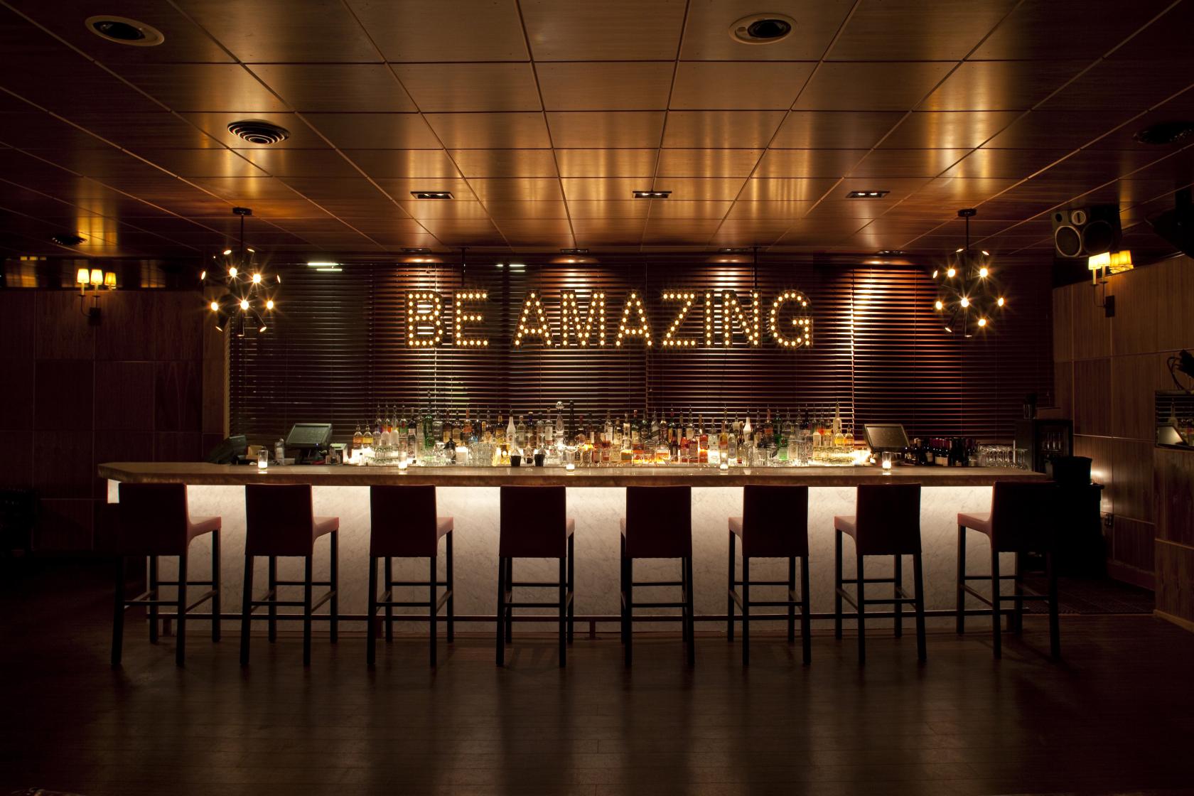 A hotel lounge with a long marble bar, backlit shelves, and marquee lights spelling 'Be Amazing' at Chambers at the Phoenix.